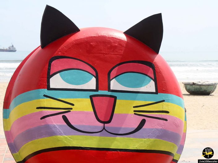 Une sculpture de chat colorée et arrondie avec des oreilles noires audacieuses, des rayures arc-en-ciel et un visage souriant est exposée à l'extérieur près d'une plage de sable et de l'océan à Da Nang au Vietnam. Un bateau et un navire sont visibles en arrière-plan.