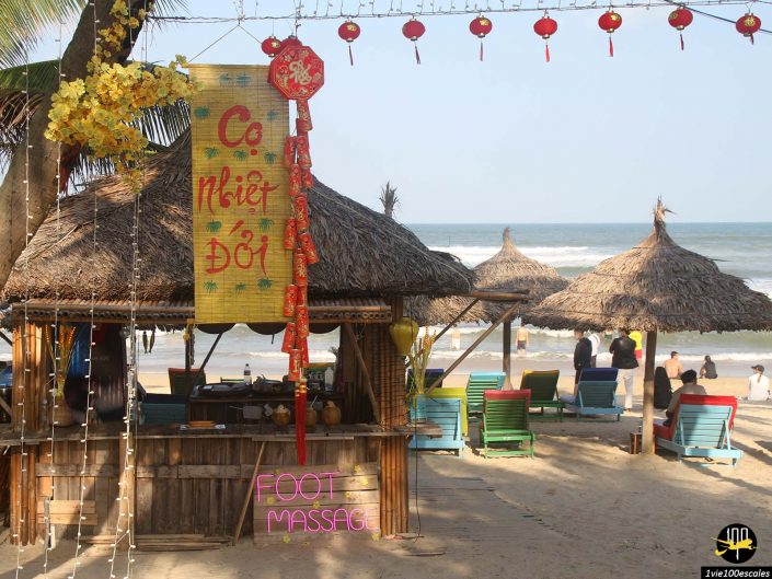 Une cabane en bord de plage à Da Nang au Vietnam propose des massages des pieds, ornée d'une pancarte « Co Nhiet Doi ». Plusieurs parasols et chaises longues au toit de chaume sont placés à proximité, avec des gens se relaxant au bord de l'océan en arrière-plan.
