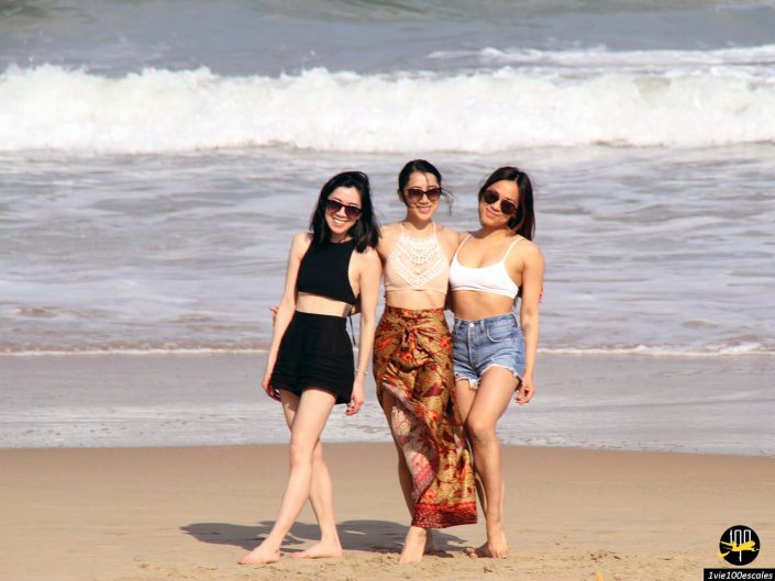 Trois femmes en tenue de plage se tiennent ensemble sur le rivage sablonneux avec les vagues de l'océan en arrière-plan, à Da Nang au Vietnam.
