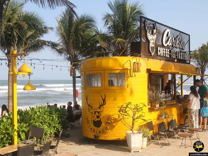 Une remorque de café jaune avec une signalisation « Caribou Coffee, Tea, Fastfood » est située près d'une plage à Da Nang au Vietnam, entourée de palmiers et de clients.