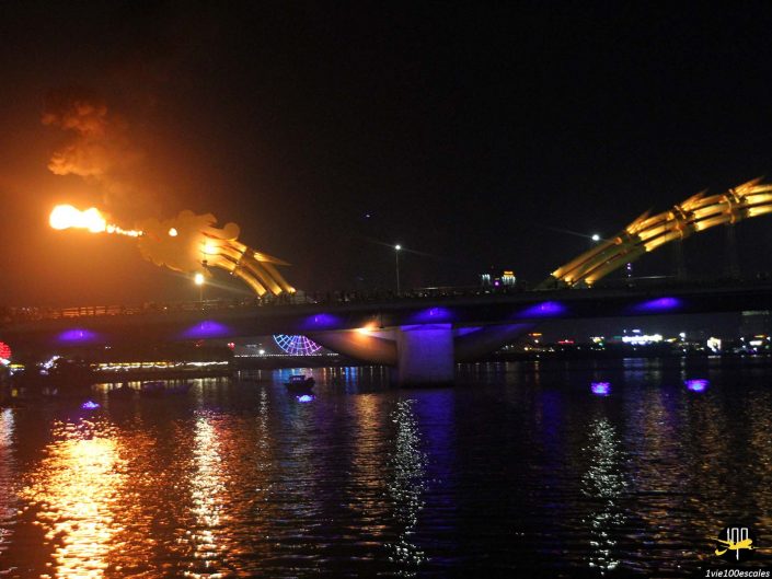 Un pont illuminé la nuit avec une structure de dragon crachant du feu au-dessus d'une rivière à Da Nang, au Vietnam. L'eau reflète les lumières et le feu.