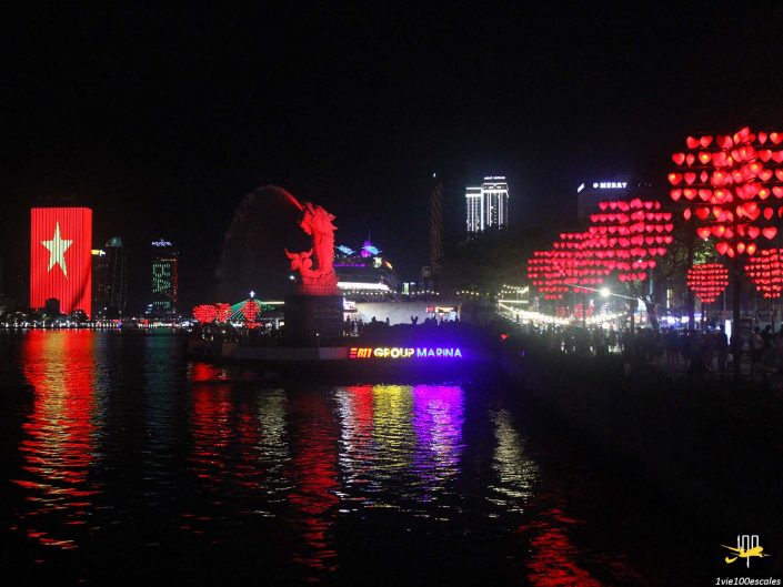 Paysage urbain nocturne présentant un front de mer à Da Nang, au Vietnam, avec des bâtiments et des décorations illuminés, une grande structure en forme d'ours émettant des lumières rouges et violettes, et une structure avec des lumières en forme de cœur à droite.