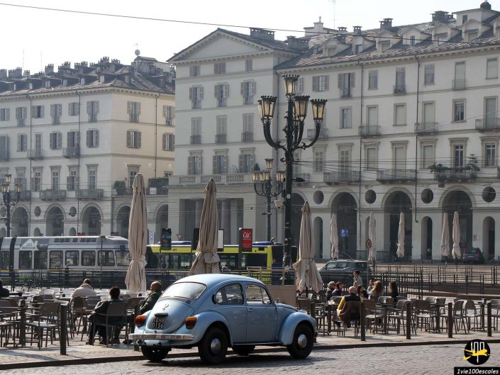 Une voiture vintage bleue est garée sur une place européenne à Turin en Italie, remplie de tables et de chaises vides. En arrière-plan, des bâtiments à plusieurs étages avec des fenêtres cintrées et des balcons. Les gens sont assis et marchent à proximité.