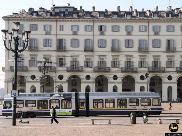 Un tramway moderne est stationné devant un élégant et grand bâtiment de couleur crème aux multiples fenêtres et arches à Turin en Italie. Le quartier semble calme avec peu de monde et un vélo à proximité.