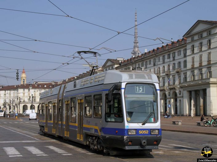 Un tramway bleu et gris portant le numéro 13 circule dans une rue d'une zone urbaine à Turin en Italie, entourée de bâtiments historiques et de caténaires.