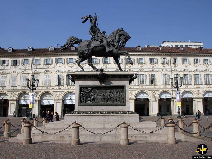 Une statue d'un cavalier à cheval lève une épée sur un piédestal devant un grand bâtiment orné de nombreuses fenêtres et portes cintrées à Turin en Italie. Des chaînes entourent la base de la statue.