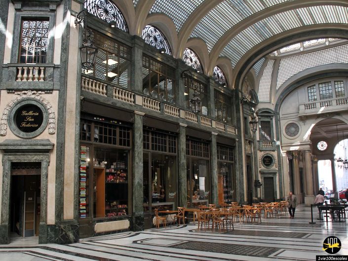 L'intérieur d'une galerie marchande ornée de hauts plafonds voûtés, de colonnes en marbre et de quelques tables à manger. Une personne marche au loin. Une plaque indique "Galleria San Federico à Turin en Italie".