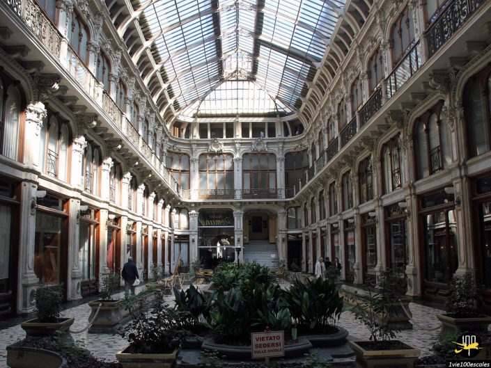 Une élégante arcade au toit de verre avec des détails architecturaux complexes abrite divers magasins et une jardinière centrale. Un panneau dans le pot indique « VIETATO SEDERSI ». Plusieurs personnes traversent ce charmant passage à Turin en Italie.