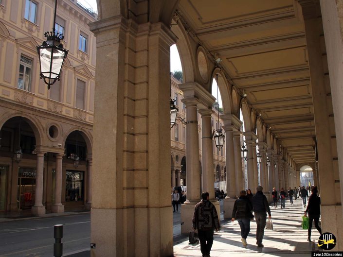 Les gens marchent sous les arcades couvertes d’un bâtiment historique avec des colonnes, avec des ombres projetées par la lumière du soleil. Le bâtiment présente des détails architecturaux élaborés et les devantures de magasins sont visibles d'un côté. Cette scène pittoresque se déroule à Turin en Italie.