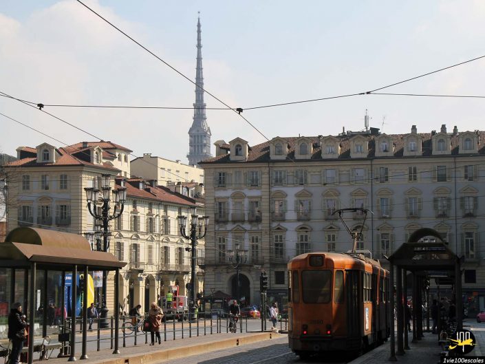 Un tramway traverse une rue bordée de bâtiments historiques, avec la flèche de la Mole Antonelliana visible en arrière-plan, à Turin en Italie.