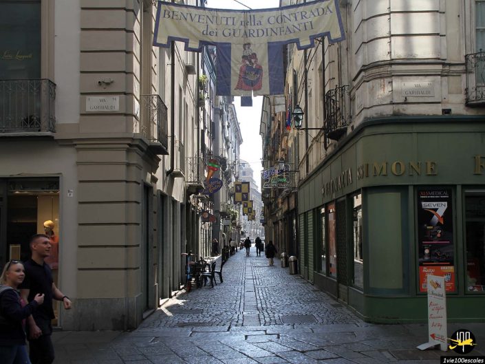 Rue pavée étroite bordée de boutiques et de cafés, de piétons au premier plan et d'une bannière au-dessus indiquant "Benvenuti Alla Contrada dei Guardinfanti" dans une ville italienne à Turin en Italie.