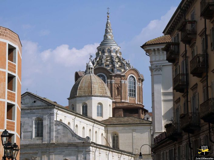 L'image montre une vue des dômes d'églises ornés et des bâtiments environnants à Turin en Italie avec un ciel clair en arrière-plan. Les structures affichent un mélange de styles et de détails architecturaux.
