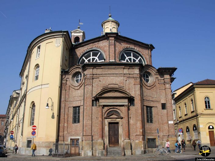 Une église en brique et en pierre avec des fenêtres circulaires se dresse sous un ciel bleu clair à Turin en Italie. A côté se trouve un bâtiment jaune. Quelques piétons et un cycliste sont visibles dans la rue devant.