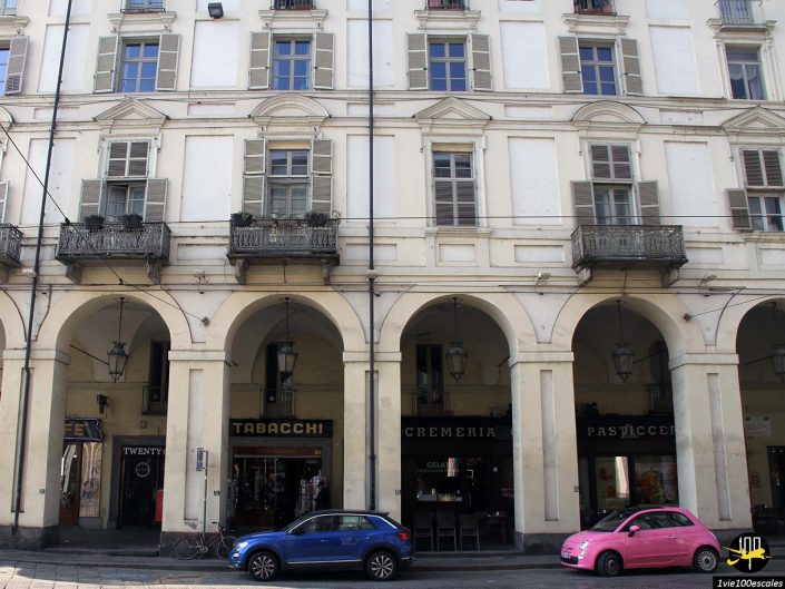 Façade d'un bâtiment historique avec fenêtres cintrées et balcons à Turin en Italie. Trois magasins se trouvent au niveau de la rue : un avec une enseigne « Tabacchi », un autre avec une enseigne « Cremeria » et un troisième avec une enseigne cyrillique. Des voitures bleues et roses sont garées devant.