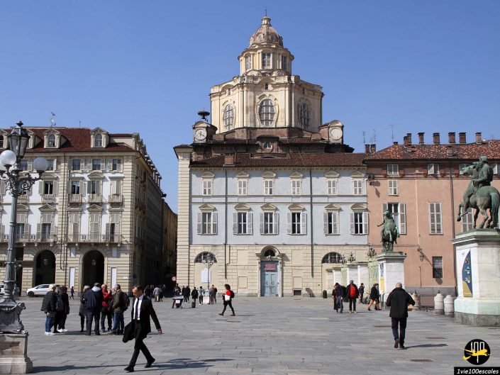 Un groupe de personnes se promène sur une place ensoleillée avec des bâtiments et des statues historiques, avec un bâtiment central orné avec un toit en forme de dôme en arrière-plan à Turin en Italie.