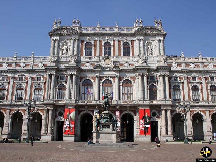 Un grand bâtiment orné de statues et de grandes fenêtres, avec une statue équestre centrale devant. Deux banderoles rouges sont accrochées de chaque côté de l’entrée. Les gens marchent dans la cour à Turin en Italie.