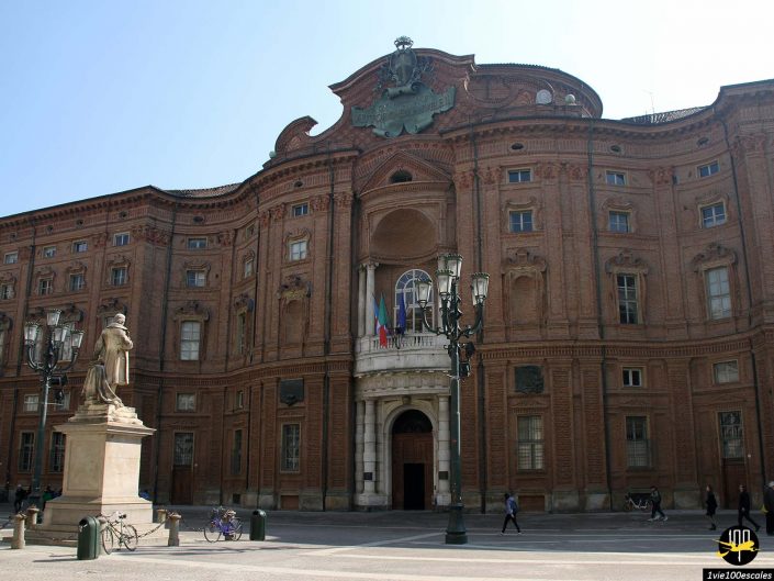 Imposant bâtiment en brique à l'architecture élaborée, doté d'un balcon central orné de drapeaux. Une statue est au premier plan et des vélos sont visibles près de l'entrée. Le ciel est clair et bleu, typique d'une belle journée à Turin en Italie.