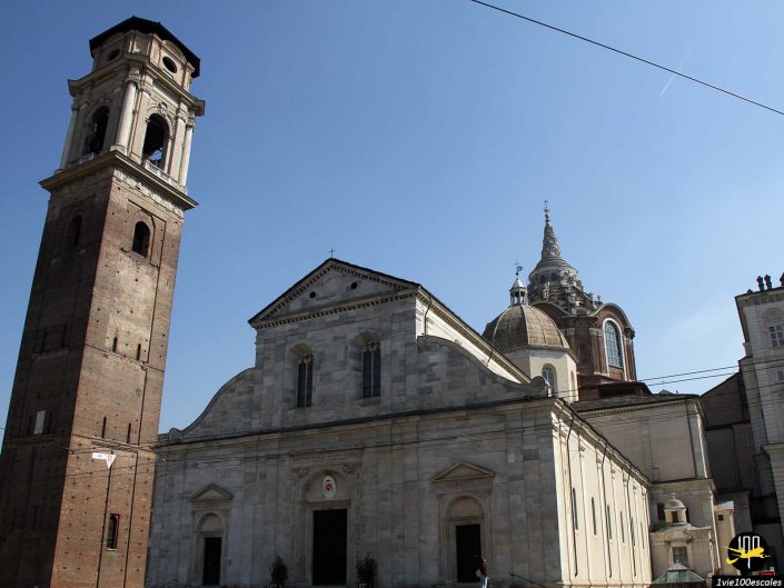 Une cathédrale en pierre avec un haut clocher en brique sur la gauche contre un ciel bleu, à Turin en Italie. La structure présente un mélange d'éléments architecturaux et de toits en forme de dôme.