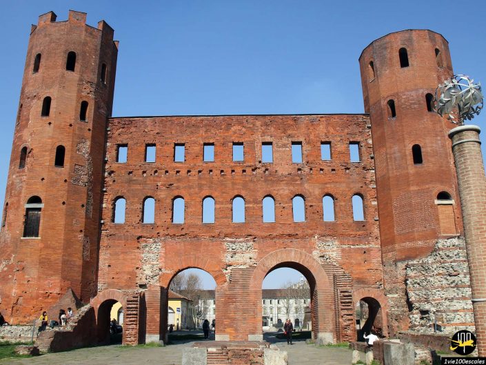 Une porte romaine historique avec deux tours circulaires, de nombreuses fenêtres rectangulaires et trois arcades se trouve en ruines à Turin en Italie, entourée de quelques éléments modernes.