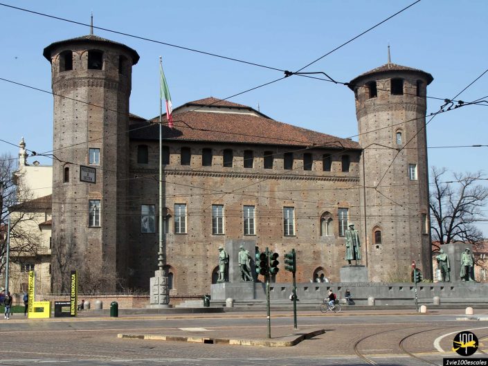Château historique en briques avec deux tours rondes, entouré de lignes de tramway et de feux tricolores. Drapeau italien bien en évidence. Quelques statues sont visibles devant la structure – à Turin en Italie.