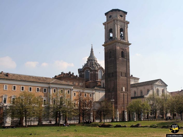 Image d’un grand clocher en brique avec un dôme pointu en arrière-plan. La tour, située à Turin en Italie, fait partie d'un complexe historique comprenant plusieurs bâtiments et une grande zone herbeuse au premier plan.