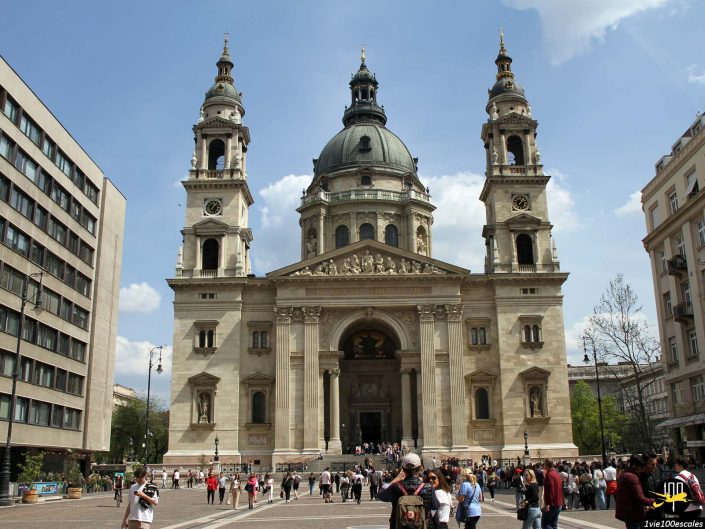Une grande cathédrale en pierre avec deux clochers et un dôme central se dresse sur une place animée remplie de monde par une journée ensoleillée, à Budapest en Hongrie.