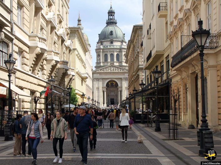 Gens marchant dans une rue pavée bordée de bâtiments historiques et de lampadaires, menant vers la basilique Saint-Étienne en arrière-plan, à Budapest en Hongrie.