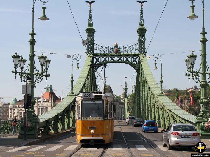 Un tramway jaune traverse le Pont de la Liberté à Budapest en Hongrie. Les voitures circulent de chaque côté du tramway et des lampadaires ornés bordent le pont. Le ciel est dégagé et les bâtiments sont visibles en arrière-plan.