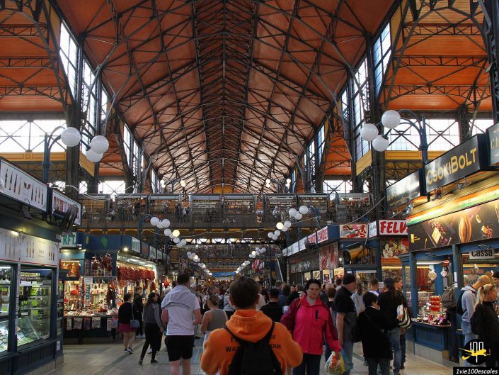Les gens marchent et font du shopping à l'intérieur d'une grande halle animée avec de hauts plafonds voûtés et de nombreux stands des deux côtés à Budapest en Hongrie.