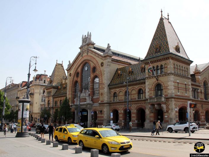 Un bâtiment de marché historique à l'architecture richement ornée et aux toits pointus se dresse dans une zone urbaine à Budapest en Hongrie, avec des taxis jaunes garés devant.