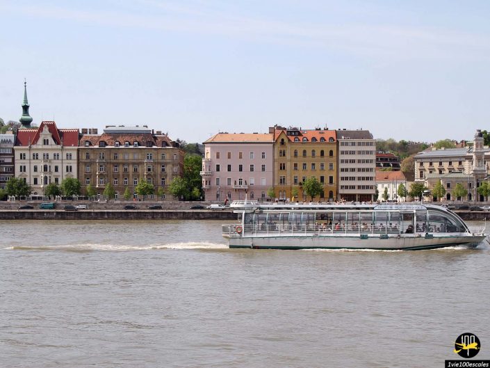 Un bateau navigue le long d'une rivière avec en toile de fond des bâtiments historiques colorés et des rues bordées d'arbres à Budapest en Hongrie.