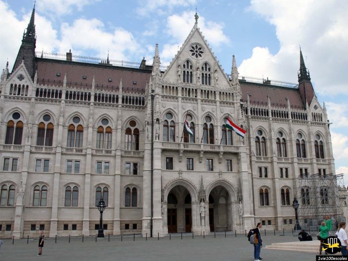 L'image montre la façade avant d'un grand bâtiment en pierre blanche orné d'éléments architecturaux gothiques à Budapest, en Hongrie, tandis que les drapeaux flottent et que les gens marchent et s'assoient au premier plan.
