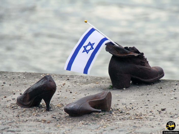 Trois chaussures en bronze, faisant partie du mémorial « Chaussures sur les rives du Danube » à Budapest en Hongrie, avec un drapeau israélien placé à l'intérieur d'une chaussure, sont présentées sur un fond flou du Danube.