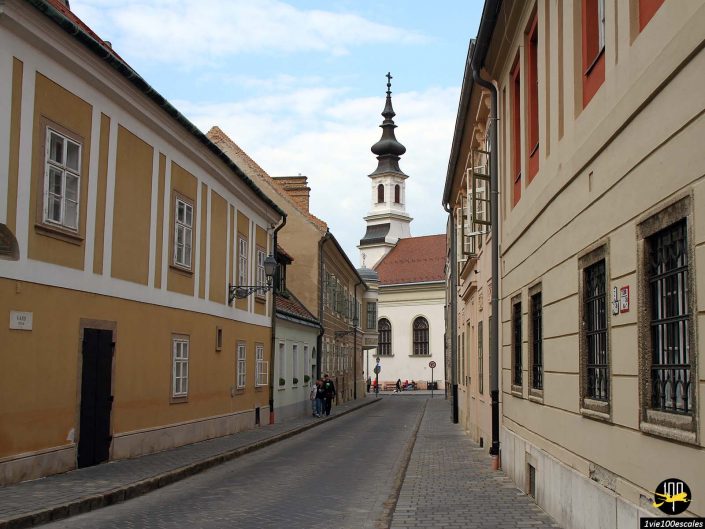 Ruelle pavée étroite avec des bâtiments historiques de chaque côté, menant à une église avec une haute flèche en arrière-plan. Quelques personnes marchent dans la rue, profitant du charme de cette scène à Budapest en Hongrie.