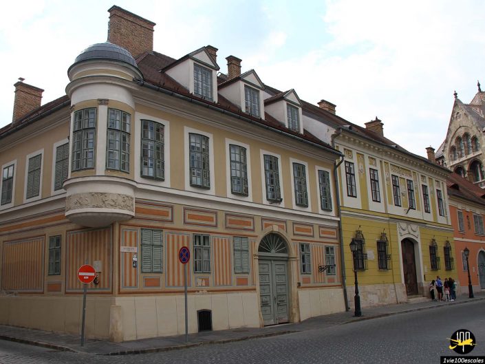 Une rangée de bâtiments historiques aux styles architecturaux variés bordent une rue pavée à Budapest en Hongrie. Un panneau d'interdiction d'entrée rouge et blanc est visible sur le côté gauche. Trois personnes marchent à proximité des immeubles.