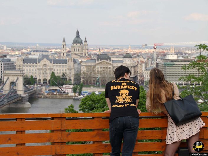 Deux personnes s'appuient sur une balustrade en bois surplombant un paysage urbain à Budapest en Hongrie, avec des bâtiments historiques importants et un pont traversant une rivière en arrière-plan.