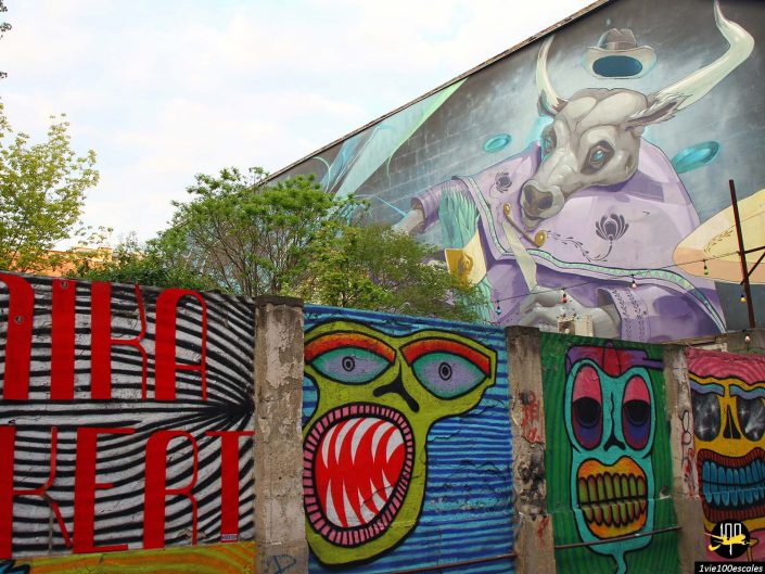 Une grande fresque représentant un taureau surréaliste en costume est peinte sur le mur d'un immeuble à Budapest en Hongrie. Au premier plan, des graffitis colorés de visages abstraits décorent une barrière, tandis que des arbres obscurcissent partiellement la vue.