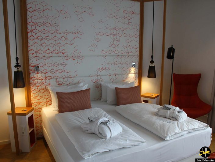 Une chambre d'hôtel moderne à Berlin en Allemagne avec un lit double, une literie blanche et deux peignoirs sur le lit. L'espace présente une décoration minimaliste, des lampes de chevet, une chaise rouge et des œuvres d'art murales géométriques derrière le lit.