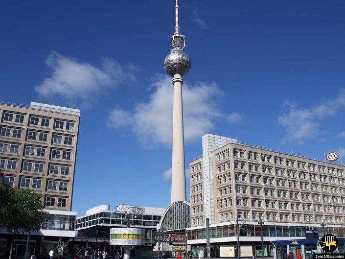 L'image montre la tour de télévision de Berlin (Fernsehturm) située sur l'Alexanderplatz à Berlin en Allemagne, entourée de bâtiments modernes et d'un ciel bleu clair.