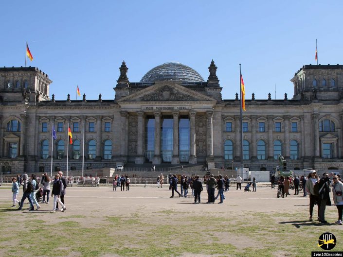 Un grand bâtiment historique avec un dôme de verre au sommet, plusieurs drapeaux et des gens marchant et se rassemblant par une journée ensoleillée à Berlin en Allemagne.