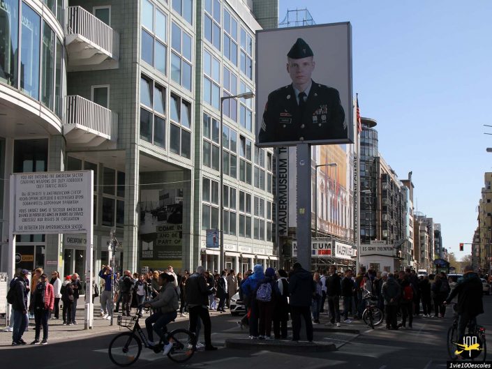 Une foule de gens dans une rue animée de la ville de Berlin en Allemagne près d'un grand panneau publicitaire affichant une personne en uniforme militaire. Des immeubles contemporains bordent la rue et divers panneaux sont visibles.