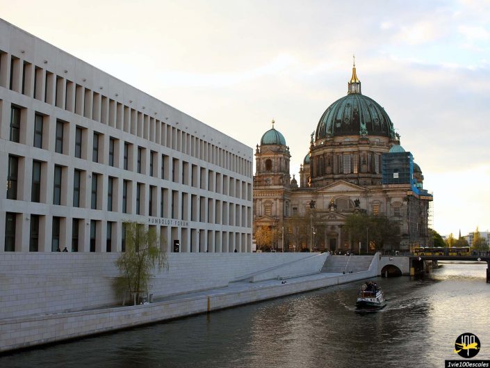 Une rivière devant un bâtiment blanc moderne et une cathédrale historique en forme de dôme avec un bateau sur l'eau, dans un paysage urbain au coucher du soleil, à Berlin en Allemagne.