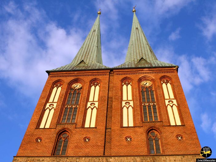Image d’une église en brique avec deux hautes flèches pointues sur un ciel bleu avec des nuages blancs. La façade présente de grandes fenêtres cintrées et des éléments décoratifs. Cette superbe pièce d'architecture est située à Berlin en Allemagne.
