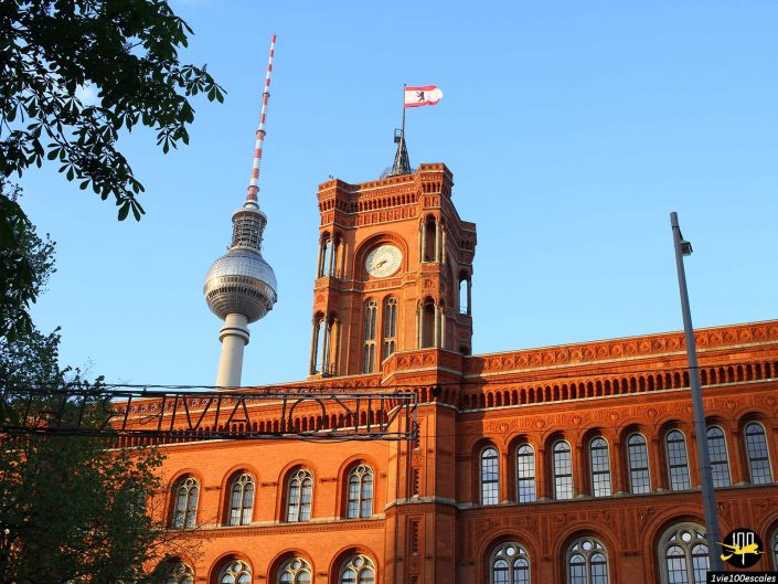 Un bâtiment historique en briques rouges surmonté d'une tour d'horloge et d'un drapeau se dresse devant une tour de télécommunications moderne, à Berlin en Allemagne. Des arbres et une structure métallique sont visibles au premier plan.