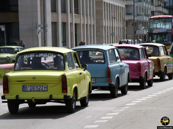Une file de voitures Trabant vintage de différentes couleurs circule dans une rue urbaine, avec des bâtiments modernes en arrière-plan, à Berlin en Allemagne.