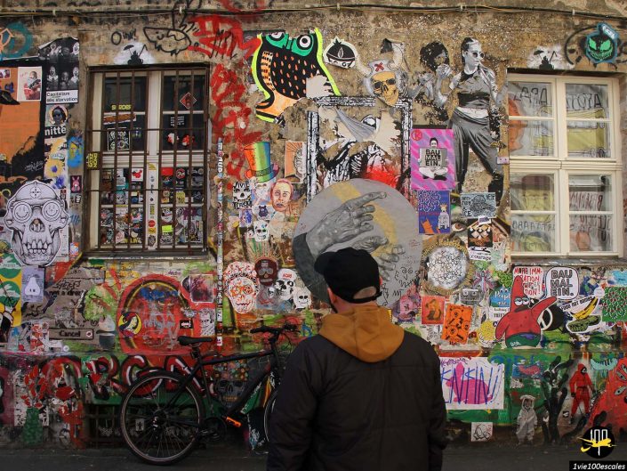 Une personne portant une veste sombre et une casquette se tient devant un mur couvert de graffitis à Berlin en Allemagne, observant les différentes œuvres d'art, notamment des peintures murales, des autocollants et des affiches. Un vélo est appuyé contre le mur.