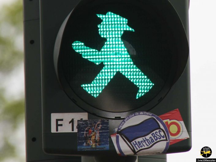 Gros plan d'un feu de circulation pour piétons à Berlin, en Allemagne, montrant une silhouette verte marchant avec un chapeau. La lumière est fixée sur un poteau orné de divers autocollants, dont un sur lequel est écrit "Hertha BSC".