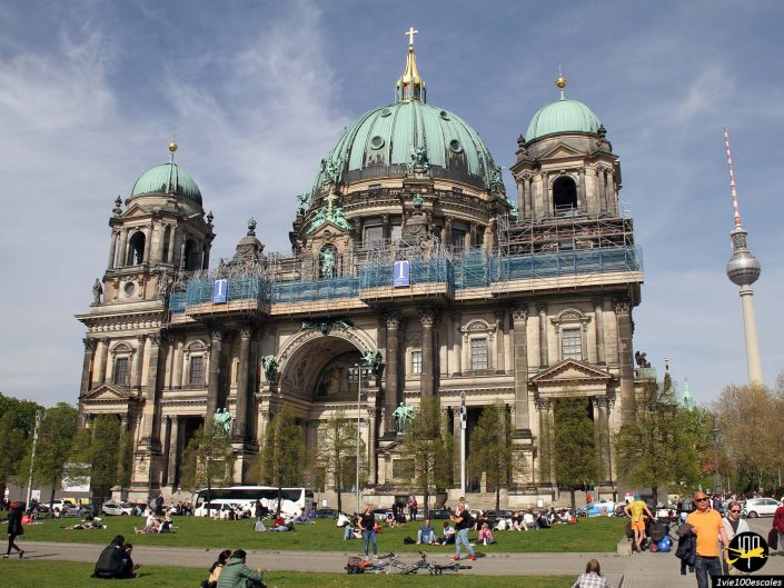 Une grande cathédrale avec des dômes verts et des échafaudages est entourée de gens qui se détendent sur l'herbe devant elle. Une haute tour de télévision se dresse à droite en arrière-plan sous un ciel partiellement nuageux à Berlin en Allemagne.