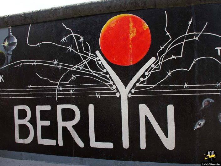 Une fresque murale avec le mot « BERLIN » en lettres grasses, avec un grand cercle rouge et des lignes artistiques ressemblant à des branches d'arbre au-dessus. L'image comprend un logo avec « 100 » et un texte qui semble dire « East Side Gallery à Berlin en Allemagne.