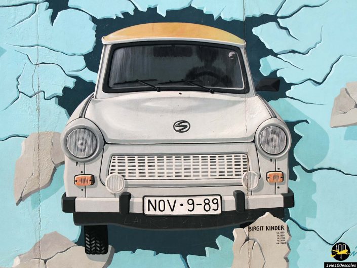 Une fresque murale à Berlin, en Allemagne, représente une voiture blanche avec un toit jaune traversant un mur. La plaque d'immatriculation de la voiture indique "NOV-9-89". L'œuvre est signée par Birgit Kinder.